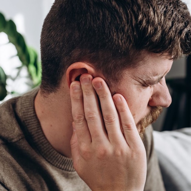Close-up of unhealthy young caucasian man 30s touching ear, suffering from sudden throbbing ear ache, looking aside. Upset bearded male feeling unwell sitting on couch in living room at home Trigeminal Neuralgia
